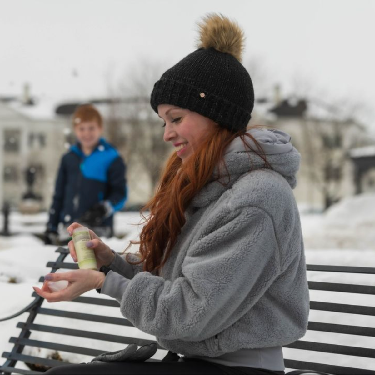 Woman sitting on a bench in a snowy park, wearing a gray jacket and black knit hat with a fur pom-pom.