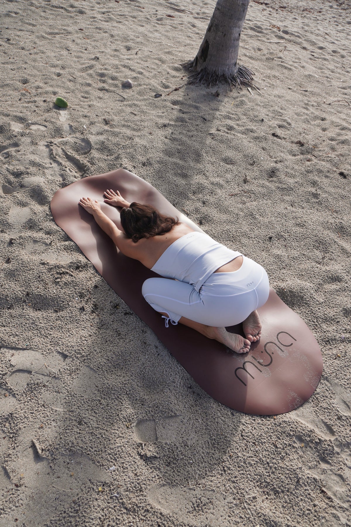 A woman in white activewear poses on a large, dark brown yoga mat placed on sandy ground, with a palm tree trunk visible on the right side.