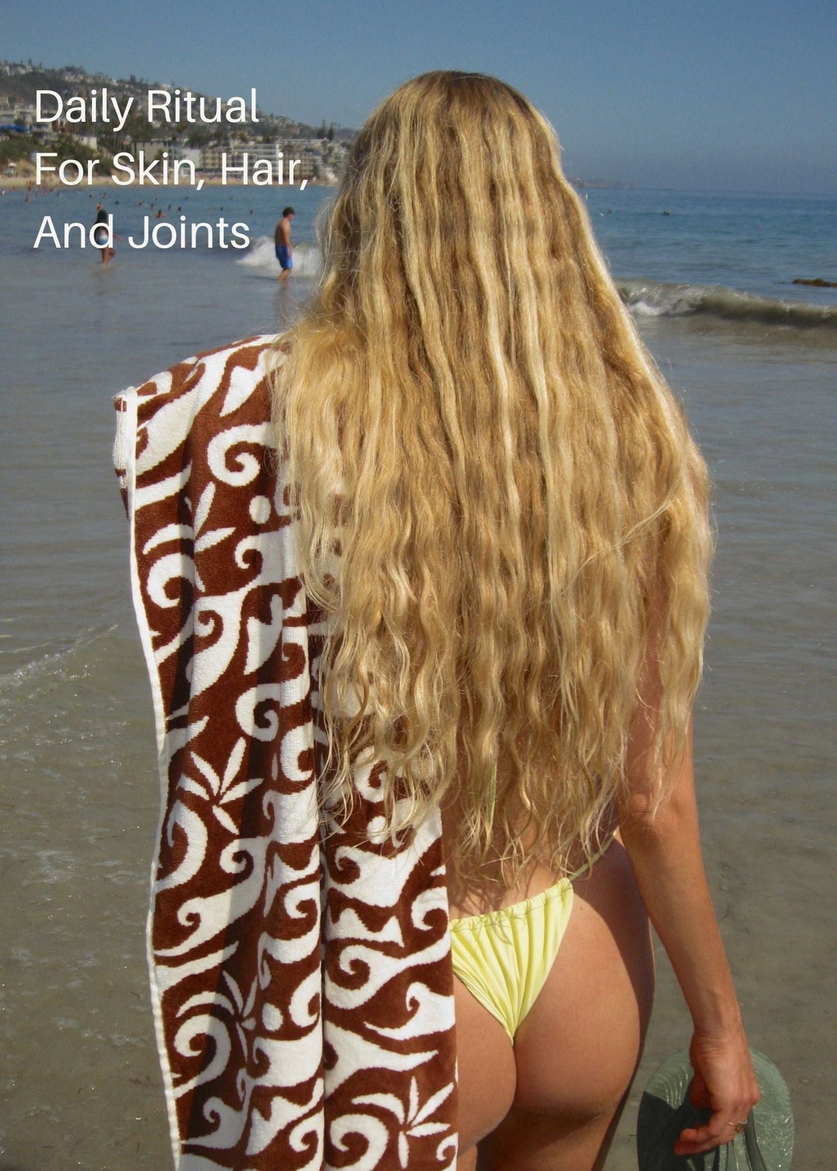 A woman with very long, wavy blonde hair stands on a beach facing the ocean, holding a brown and white patterned towel over her shoulder. She is wearing a yellow bikini bottom.