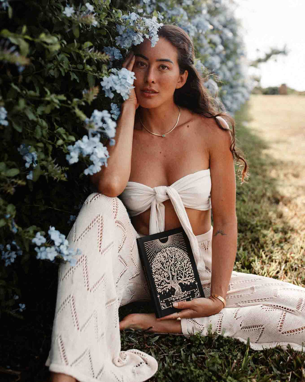 Woman in a white outfit holding a book in a natural setting with flowers and greenery.