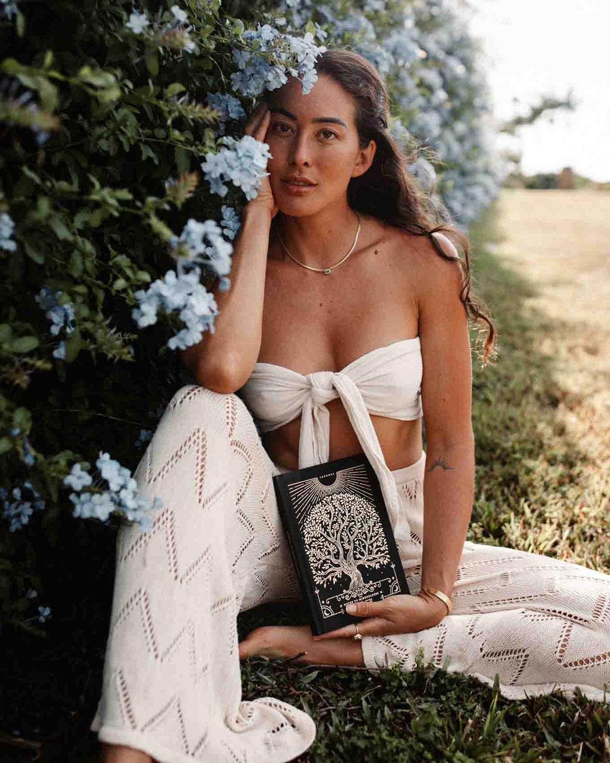 Woman in a white outfit holding a book in a natural setting with flowers and greenery.