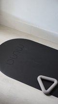 A black oval yoga/exercise mat with the brand name 'minu' debossed on it, shown on a light wood floor next to a gray foam handle.