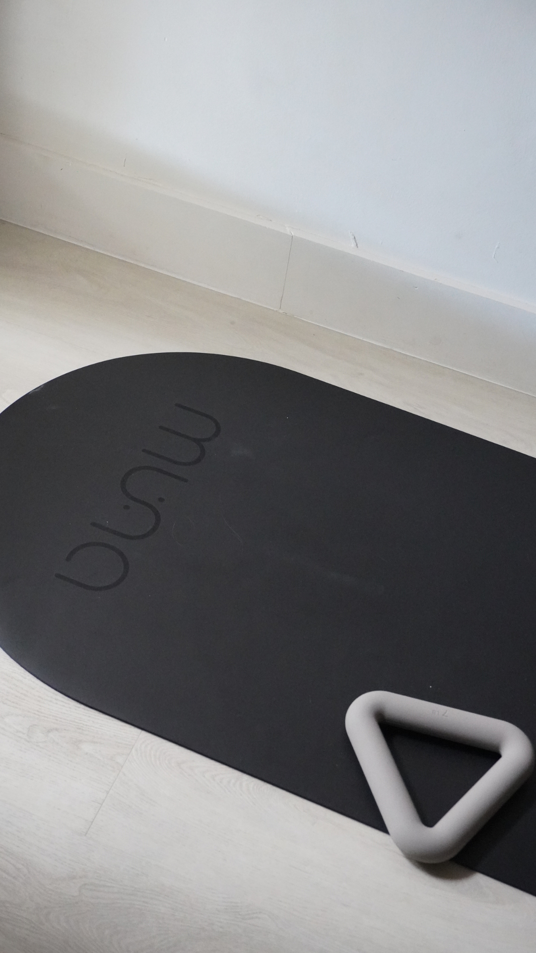 A black oval yoga/exercise mat with the brand name 'minu' debossed on it, shown on a light wood floor next to a gray foam handle.