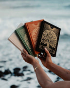 Three books with decorative covers held by a person against a water background