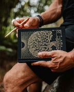 Person holding a black book with gold tree design and a pen outdoors