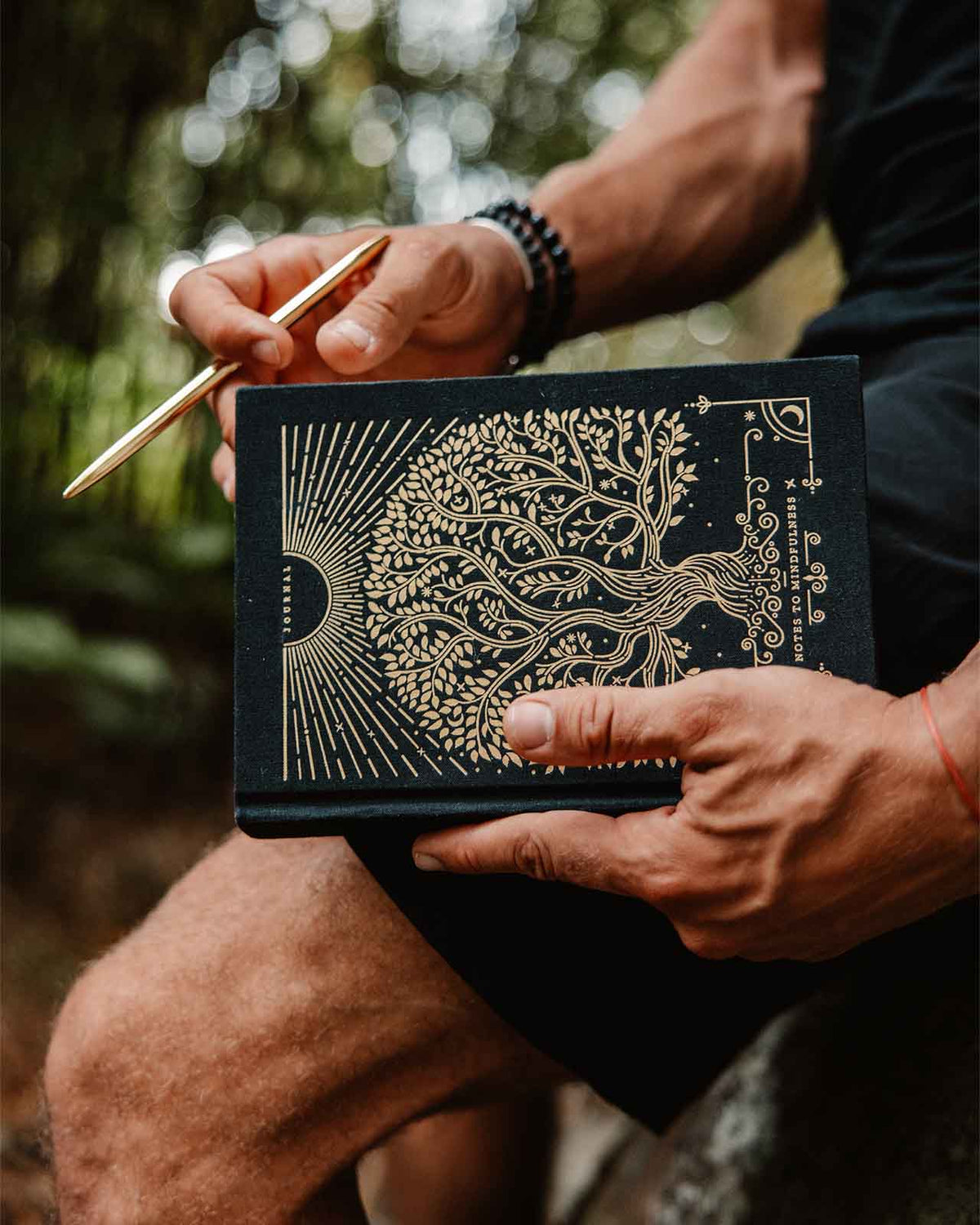 Person holding a black book with gold tree design and a pen outdoors