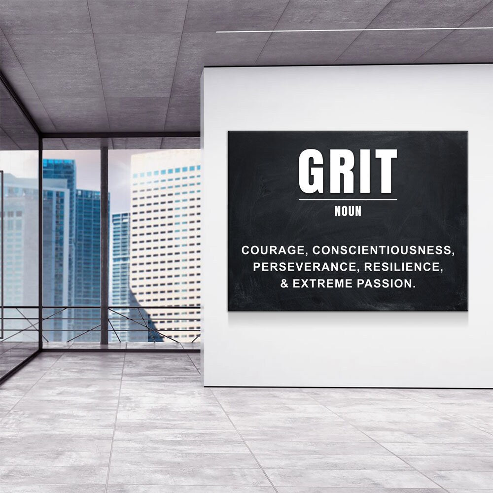 A black rectangular wall art print with white text reading 'GRIT' and a dictionary-style definition, displayed on a white wall in a modern office interior with large windows and city buildings outside.