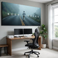 A large rectangular wall art print hangs above a wooden desk in a modern home office. The artwork shows a person walking on a foggy road with the words 'KEEP GOING' across it.