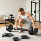 A man in a white tank top and gray shorts is crouching on a light rug while holding a black adjustable dumbbell. Several black weight plates are scattered around him on a light-colored floor in a bright home gym setting.