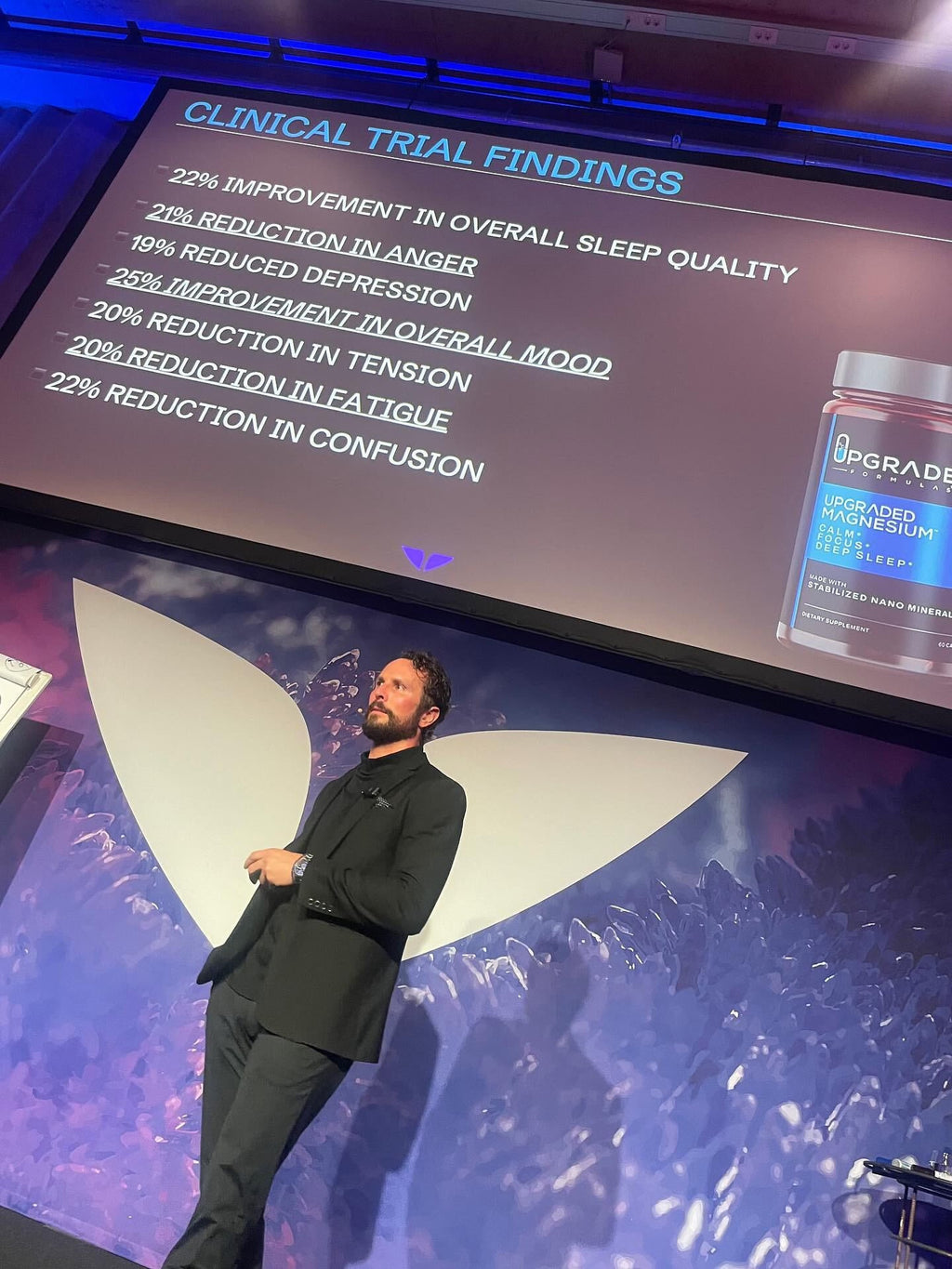 Man standing on a stage with a screen displaying clinical trial findings and a bottle of 'Upgrade' supplement.