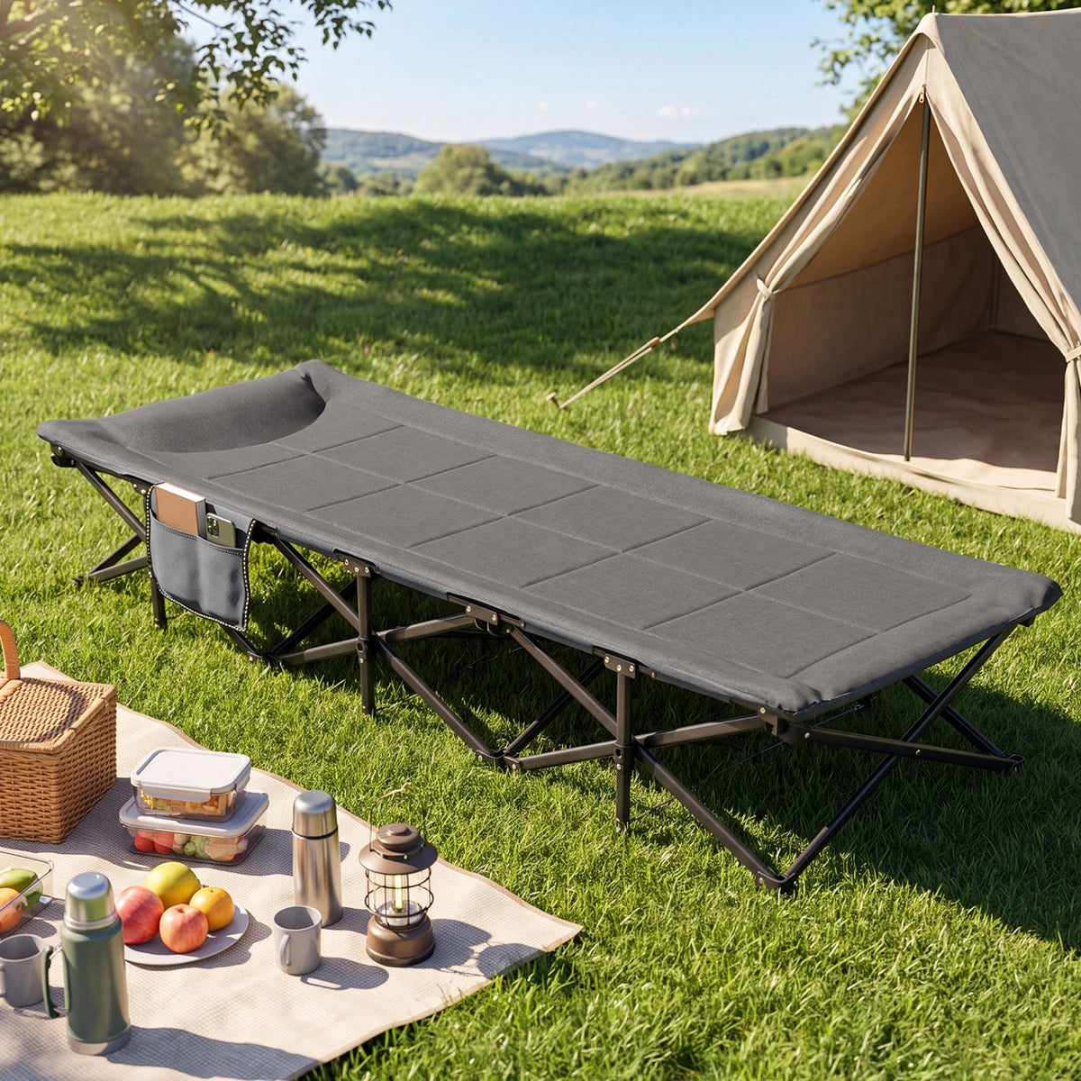 A dark gray folding camping cot with a side storage pocket, set up on a green grassy lawn next to a canvas tent and a picnic blanket with outdoor gear.