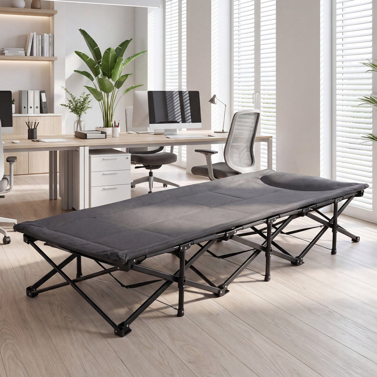 A dark gray portable camping cot with a black metal frame, shown set up in a bright, modern home office environment with wooden floors, desks, and office chairs.