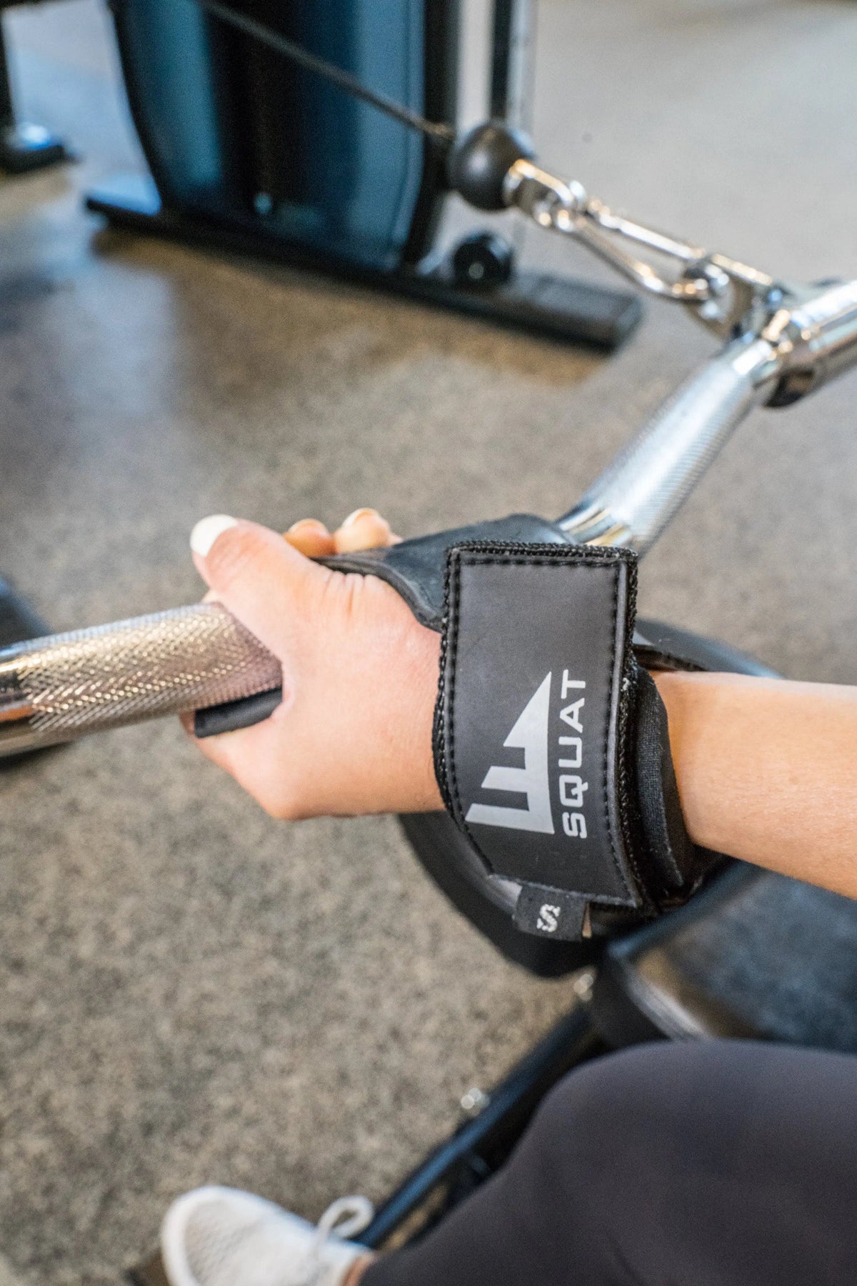 A close-up of a person's hand gripping a cable pulley machine handle in a gym, wearing a black wrist strap with the brand name SQUAT printed in white.