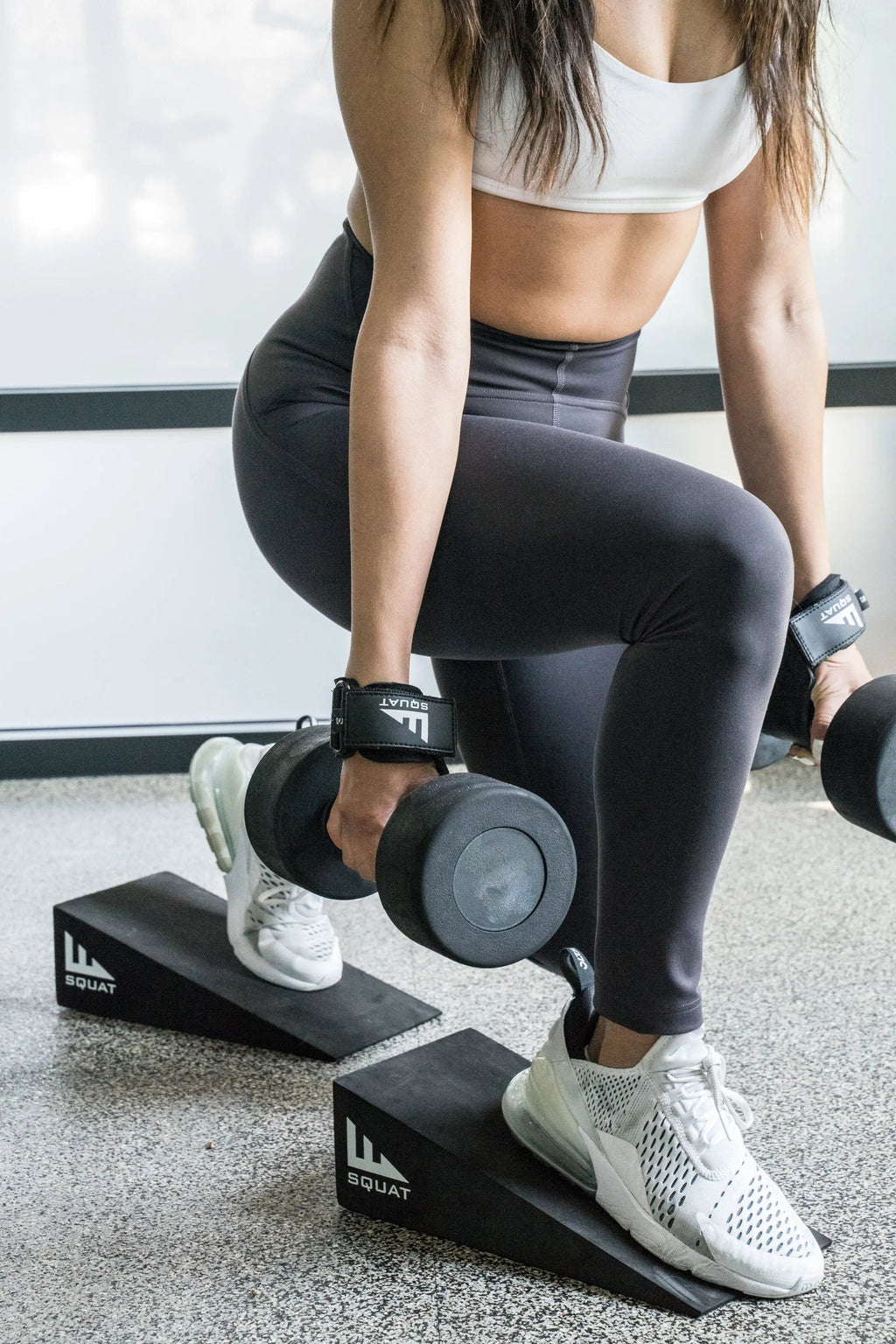 A woman in a white sports bra and black leggings performs a squat using dumbbells and black SquatWells weight platforms in a bright gym setting.