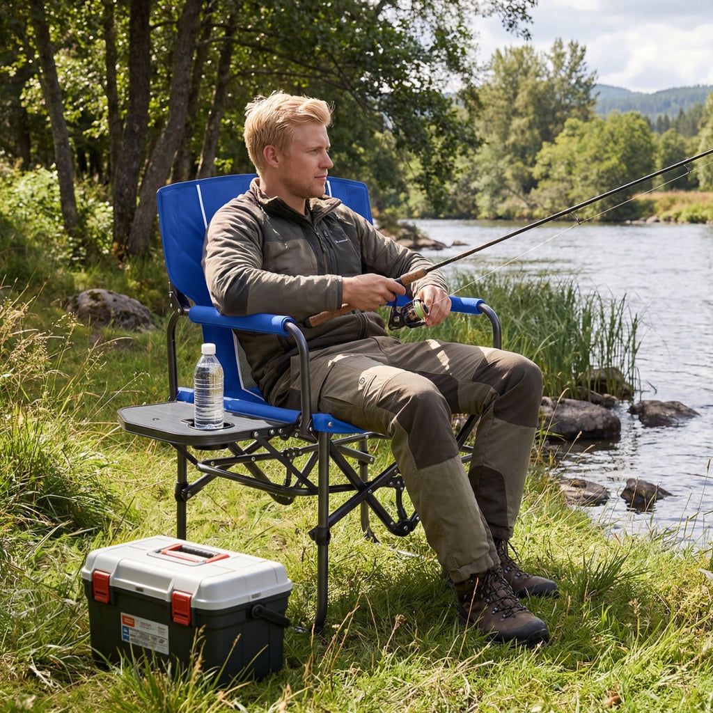 A man sits in a blue folding camp chair by a river, holding a fishing rod. The chair has a small side table with a water bottle and a black-and-white cooler on the grass nearby.