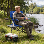 A man sits in a blue folding camp chair by a river, holding a fishing rod. The chair has a small side table with a water bottle and a black-and-white cooler on the grass nearby.