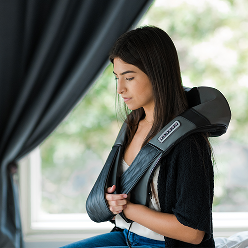 Woman using a black and gray massage device on her back in a room with a window.