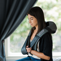 Woman using a black and gray massage device on her back in a room with a window.