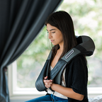 Woman using a black and gray massage device on her back in a room with a window.