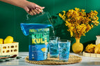 Person pouring blue Kuli Kuli powder into glasses with ice on a table with yellow flowers and lemons.