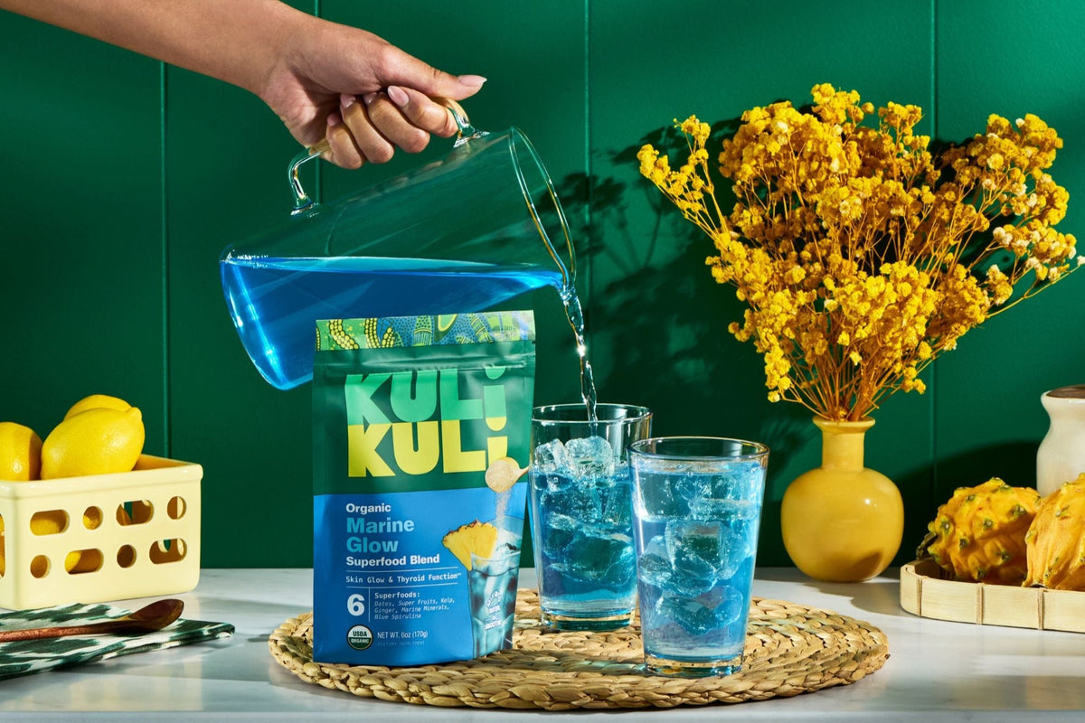 Person pouring blue Kuli Kuli powder into glasses with ice on a table with yellow flowers and lemons.