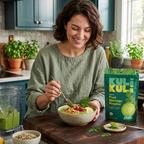 A woman in a green shirt stands in a kitchen, smiling while eating from a bowl of green smoothie. A stand-up pouch of Kuli Kuli Pure Moringa Powder sits on the counter beside her, along with a spoon, a small dish of green powder, and a blender jar containing the green drink.