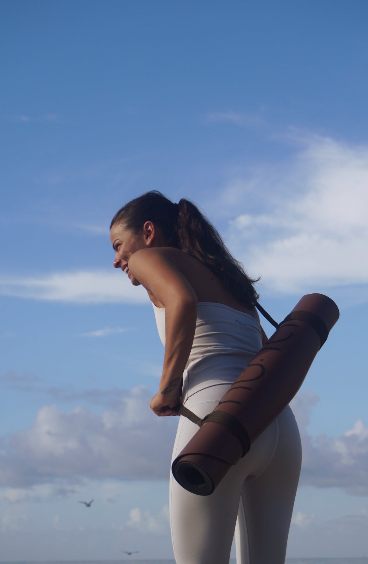A woman in a white sports bra and leggings holds a rolled brown yoga mat against her back, set against a bright blue sky with light clouds.