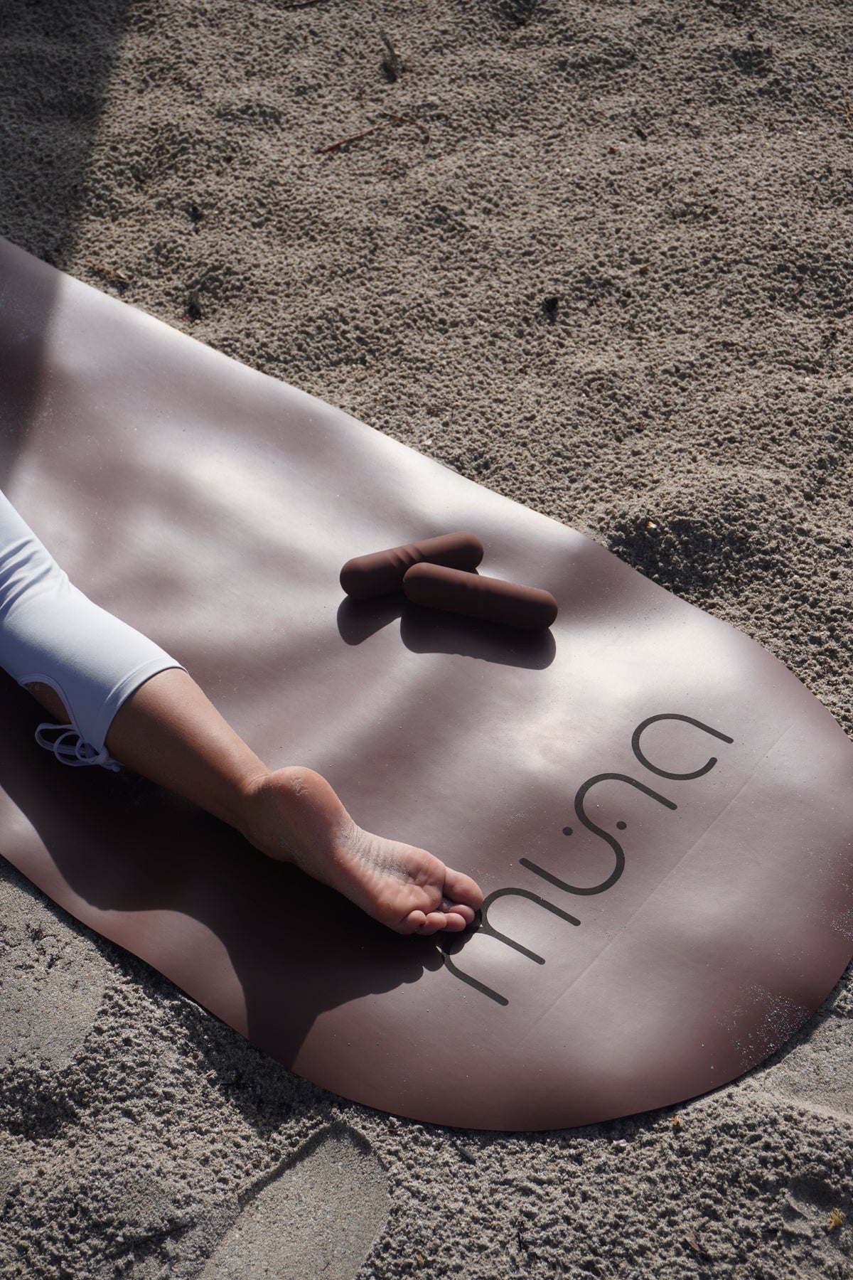 A brown yoga mat with the brand name 'mina' printed in black, laid out on sandy ground. A person's leg and foot are resting on the mat, and two brown yoga props are placed nearby.
