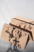 A beige foldable card case with a printed botanical shadow pattern, shown partially open on a light gray surface with dappled shadows cast across it. The brand name 'muno' is printed on the front.