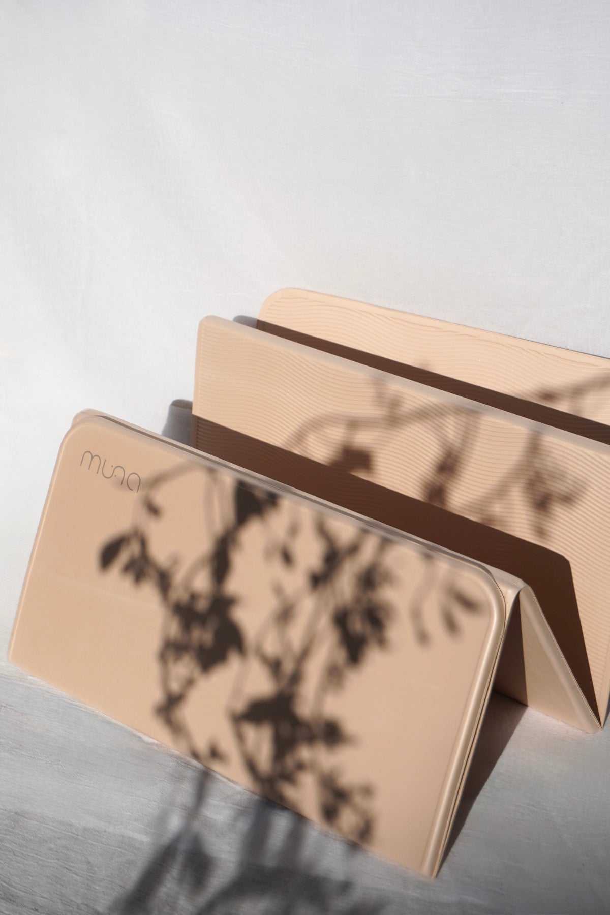 A beige foldable card case with a printed botanical shadow pattern, shown partially open on a light gray surface with dappled shadows cast across it. The brand name 'muno' is printed on the front.