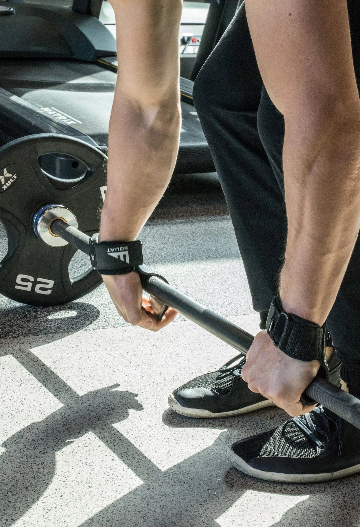 Close-up of a person lifting a barbell in a gym, wearing black wrist wraps and black athletic shoes on a concrete floor.