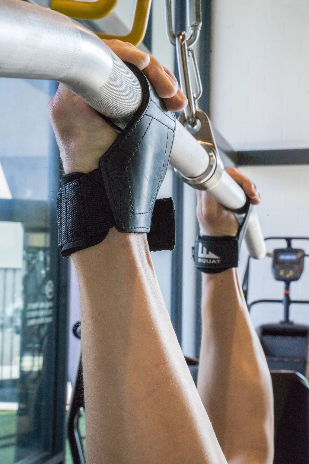 Close-up of a person's arms and hands gripping a silver pull-up bar in a gym, wearing black wrist wraps with white branding.