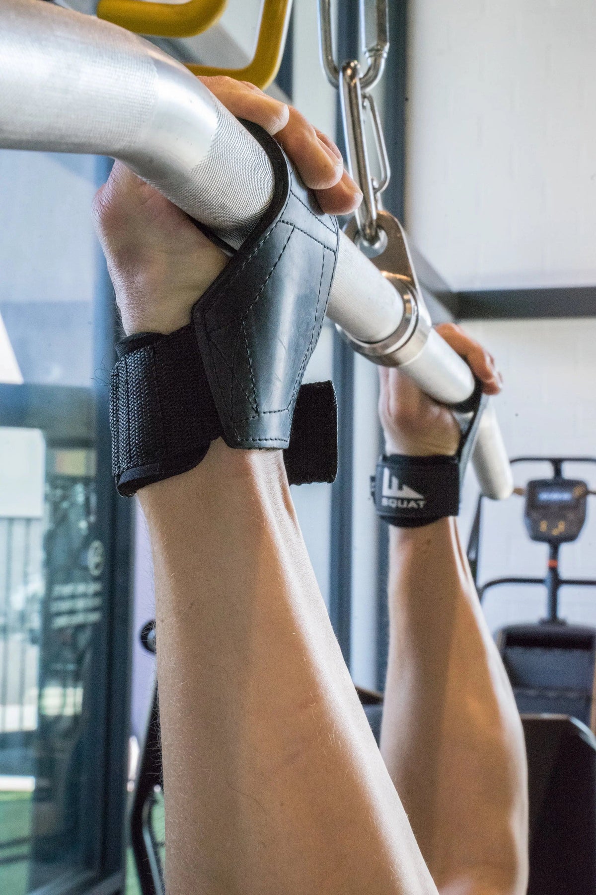 Close-up of a person's arms and hands gripping a silver pull-up bar in a gym, wearing black wrist wraps with white branding.
