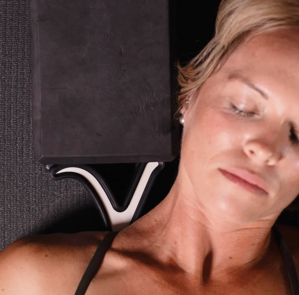 A close-up lifestyle shot of a woman lying down with her eyes closed, using a black foam meditation block on a textured dark gray mat.