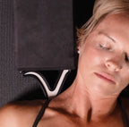 A close-up lifestyle shot of a woman lying down with her eyes closed, using a black foam meditation block on a textured dark gray mat.