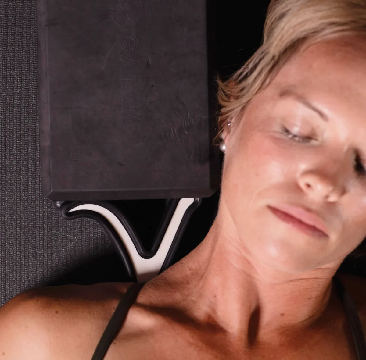 A close-up lifestyle shot of a woman lying down with her eyes closed, using a black foam meditation block on a textured dark gray mat.