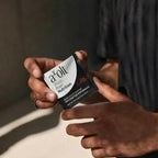 A close-up of a person holding a single aOLT Sharp Brain Nutrition supplement packet. The packet is black and white with red accents, and the person's hands are visible holding it against a dark, moody background.