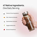 A hand holds a clear glass water bottle with a silver lid, filled with a dark brown herbal tea blend. The image includes marketing text on a light pink gradient background: '47 Native Ingredients.
