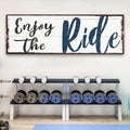 Wooden sign with 'Enjoy the Ride' text above a weight bench with dumbbells in a gym setting.