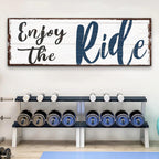 Wooden sign with 'Enjoy the Ride' text above a weight bench with dumbbells in a gym setting.