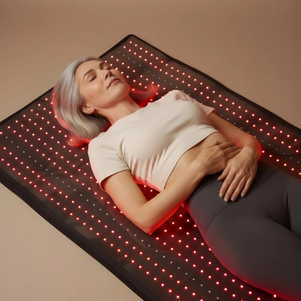 A woman lies on a black LED light therapy mat with glowing red lights, wearing a white crop top and dark gray leggings against a warm beige background.